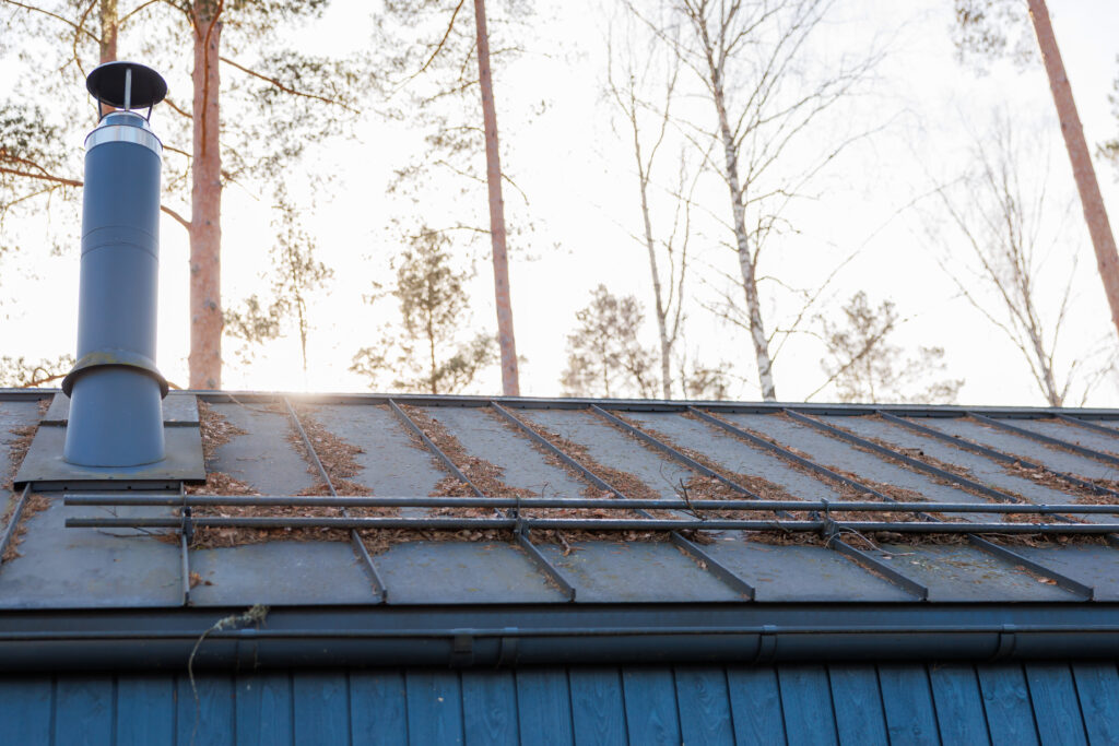 A blue painted building's standing seam metal roof holds pine needles as a chimney with rain cap rises near the ridge, snow guards span panels, soft backlighting.